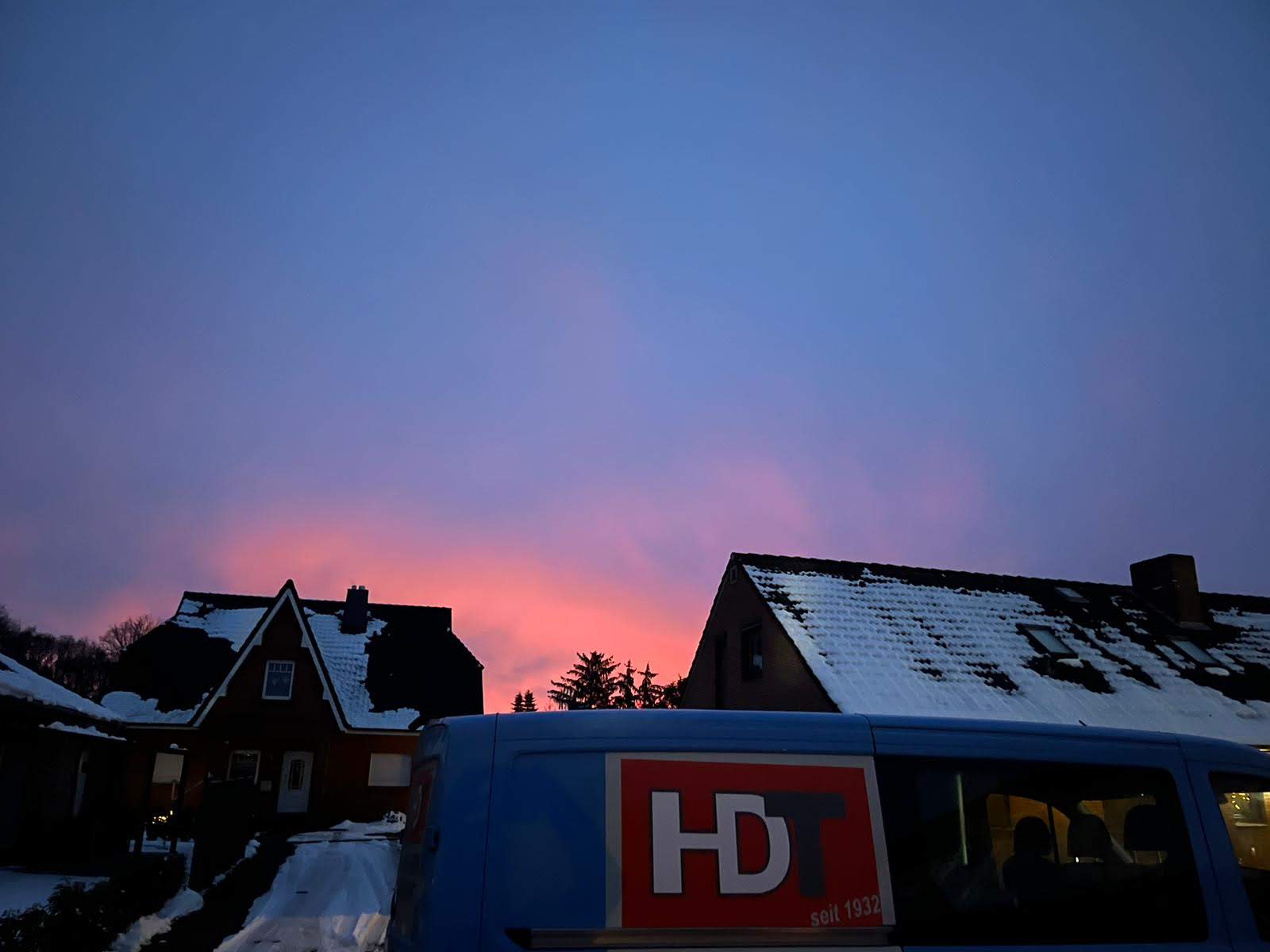 Sonstiges (16) Sonnenuntergang mit pink-blauem Himmel über verschneiten Hausdächern – im Vordergrund ein Fahrzeug mit HDT-Logo.