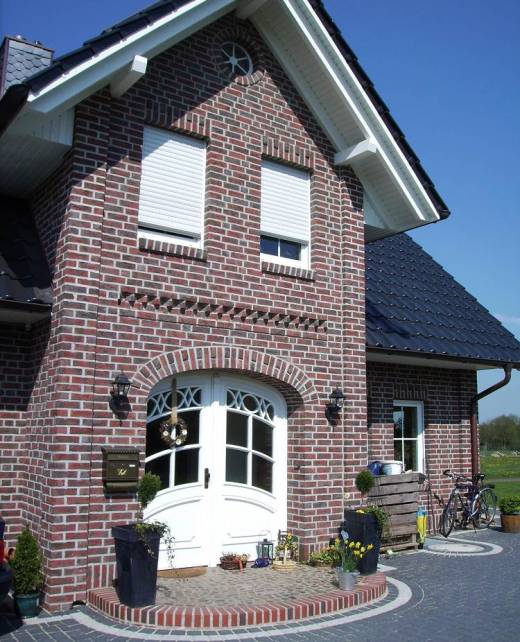 Referenzen-10 Eingangsbereich eines modernen Einfamilienhauses mit rotem Klinker, Rundbogen über der weißen Haustür und dekorativ bepflanzter Fläche.